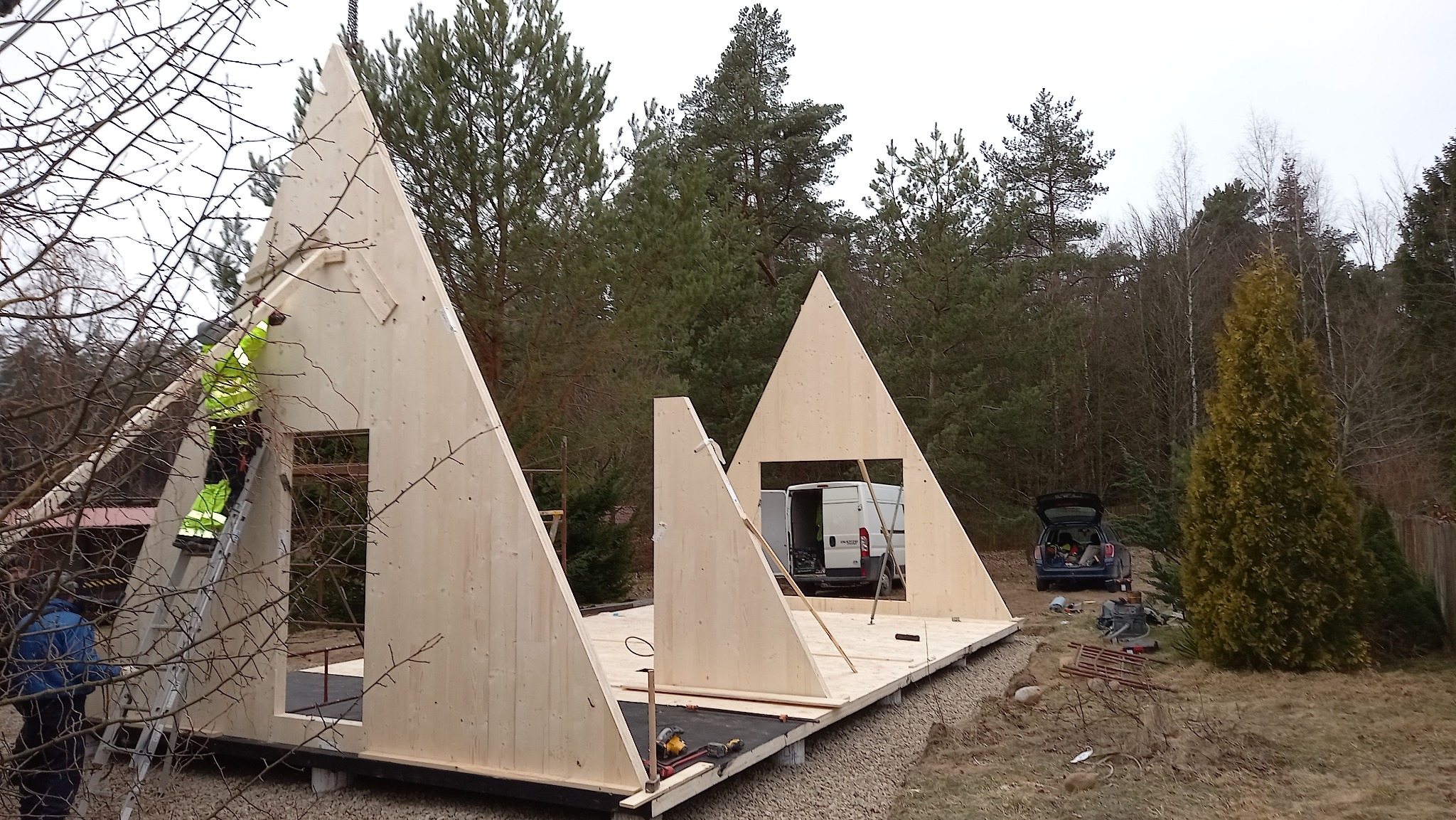 Montaż ścian szkieletowych budynku na fundamencie, widoczni pracownicy w ubraniach roboczych, na tle lasu i zaparkowanych pojazdów.