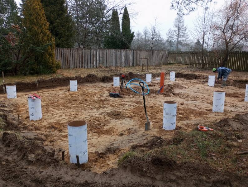 Przygotowanie terenu pod budowę: widoczne wykopane fundamenty, szalunki cylindryczne i pracownik w oddali.