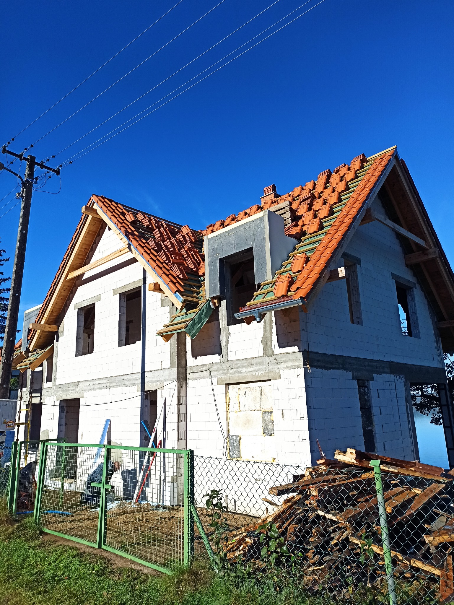 Wznoszony dwukondygnacyjny budynek z białych bloczków, częściowo pokryty czerwoną dachówką, widoczny przez zielone ogrodzenie z siatki, na tle błękitnego nieba.