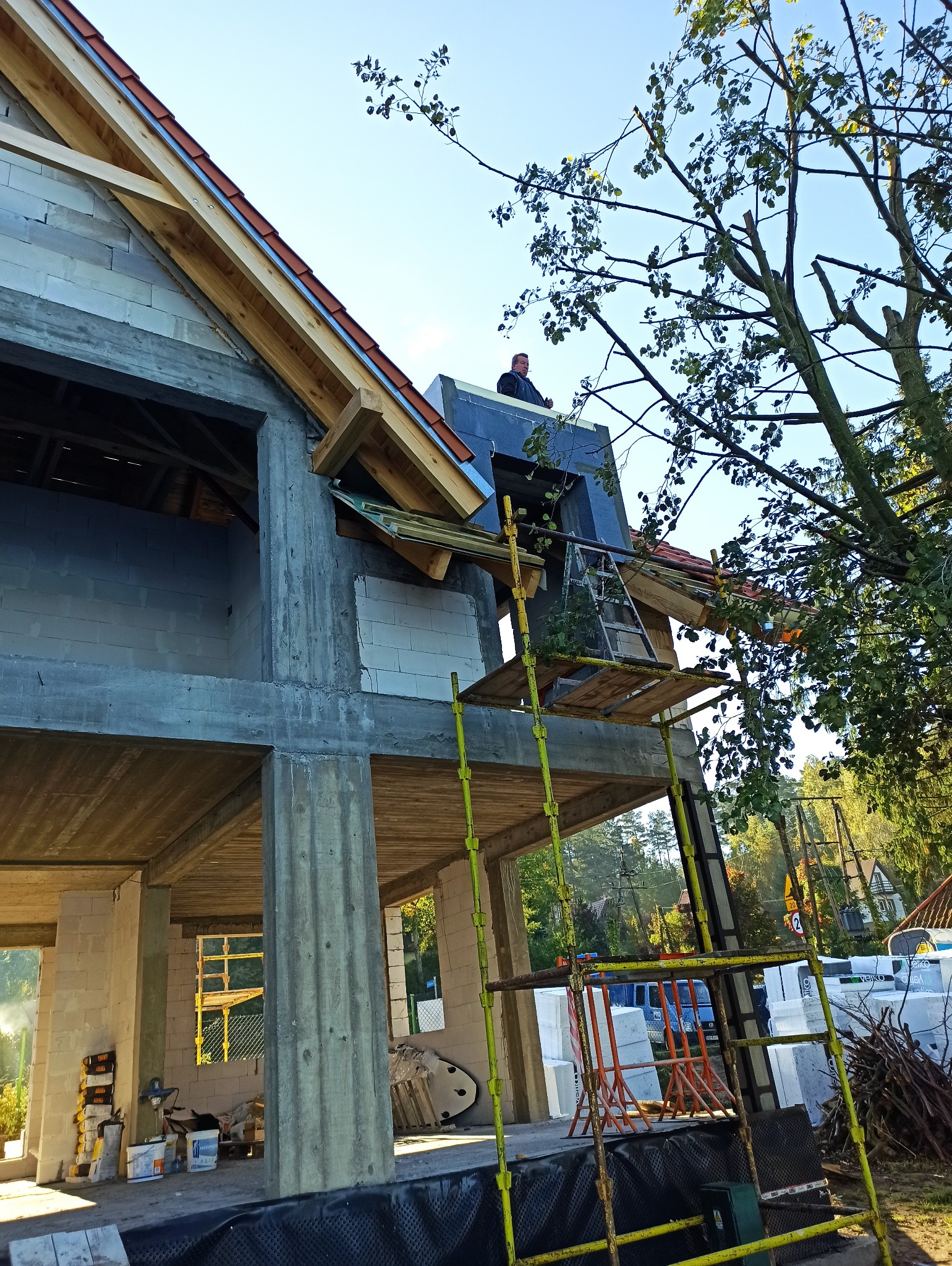 Konstrukcja domu w stanie surowym otwartym, widoczne betonowe słupy, ściany z bloczków, drewniana więźba dachowa, rusztowania i pracownik na dachu w trakcie prac dekarskich.