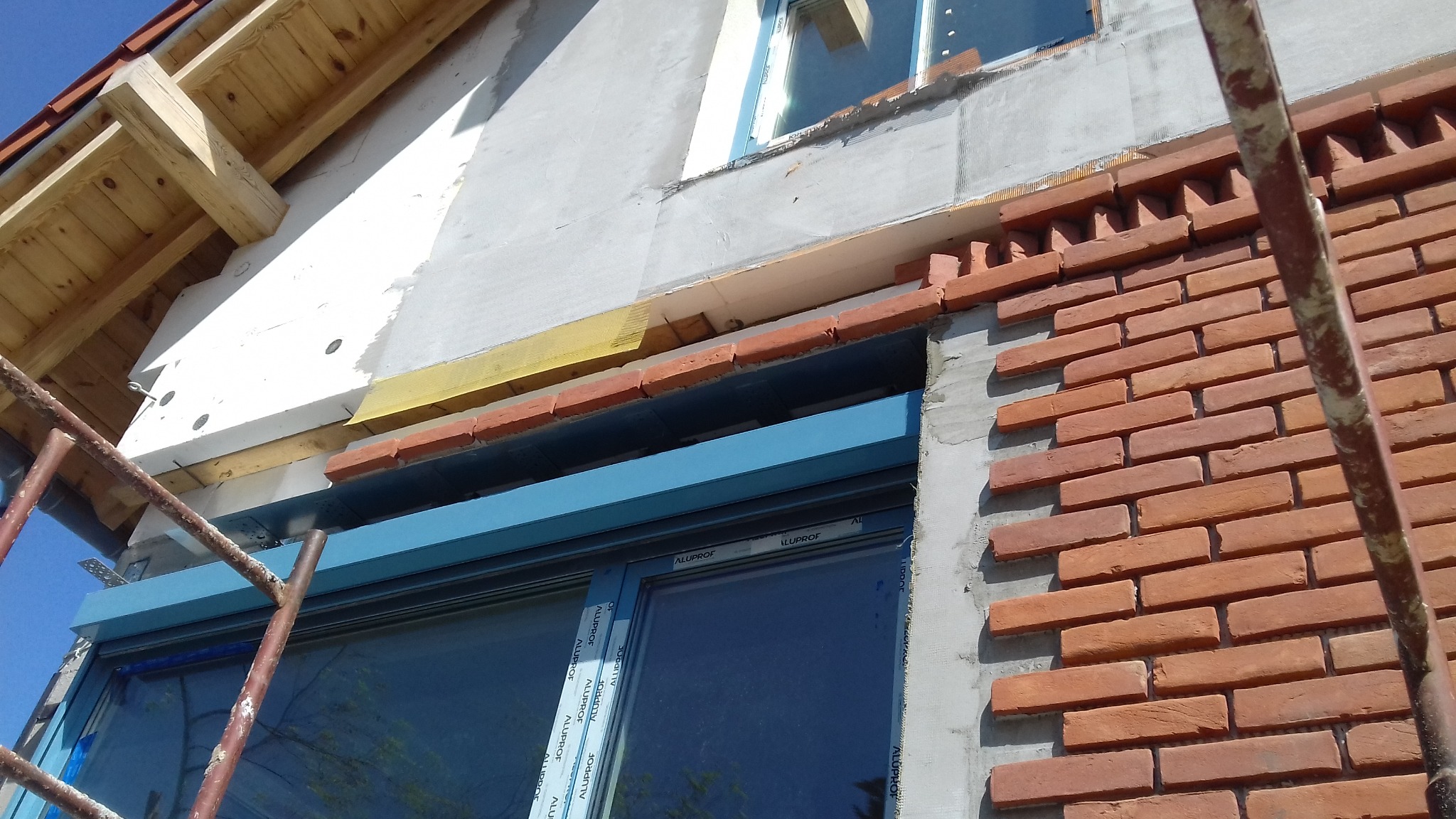 Wznoszenie muru z czerwonej cegły klinkierowej wokół okna z niebieską ramą, widoczne rusztowanie i fragment dachu z drewnianą konstrukcją, ocieplenie budynku w trakcie prac.