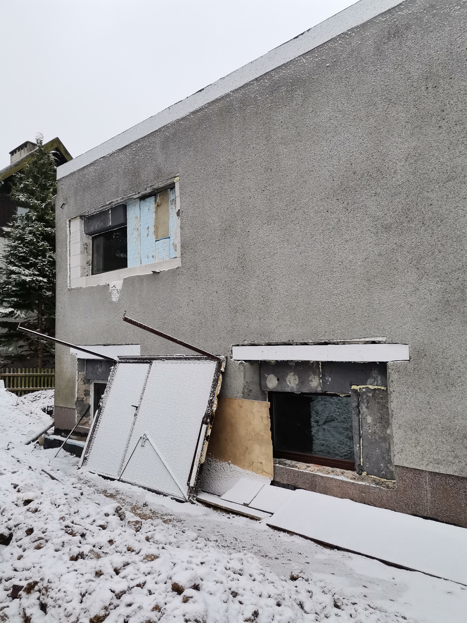 Szary budynek z odkrytymi otworami okiennymi i częściowo zdjętą bramą garażową, pokryty śniegiem, w trakcie prac remontowych lub rozbiórkowych, widoczne elementy izolacji i gruzu.