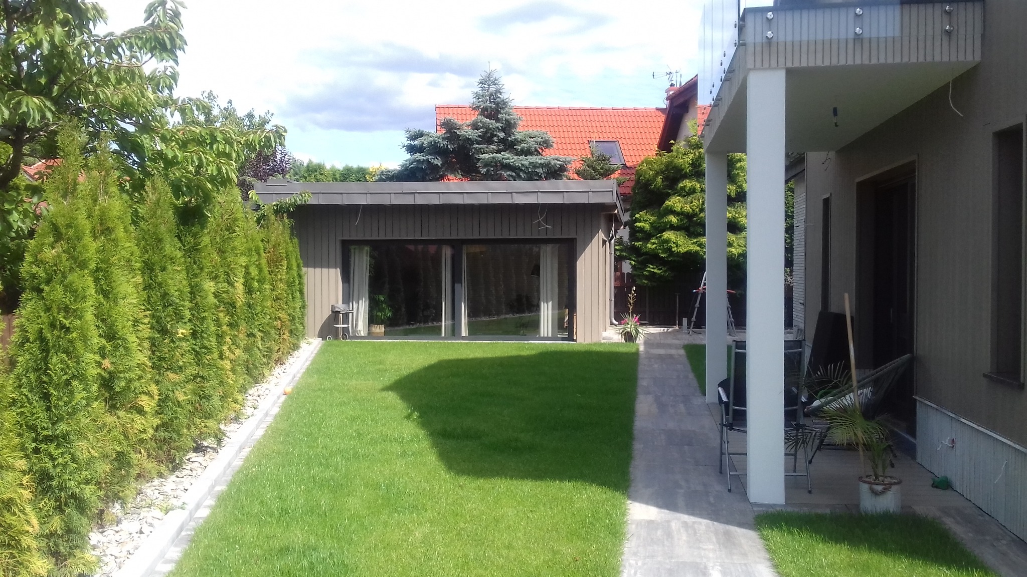 Nowoczesny parterowy dom z drewnianą elewacją i dużymi oknami przesuwnymi, otoczony zielonym trawnikiem i tujami, widok z boku budynku z zadaszonym tarasem.