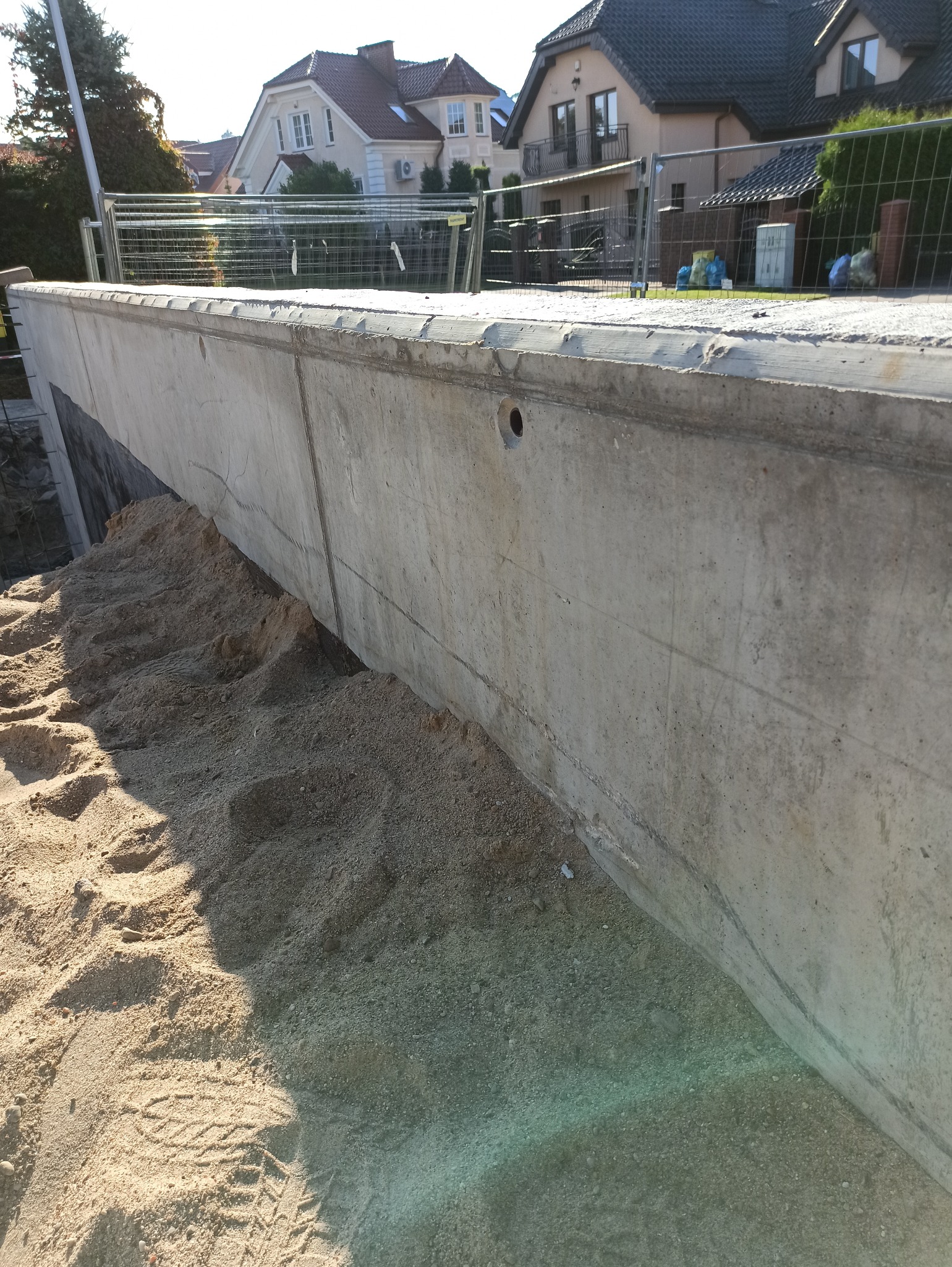 Żelbetowy fundament pod budowę domu, zasypany piaskiem, widoczne ślady opon i otwór w betonie, w tle ogrodzenie i domy.