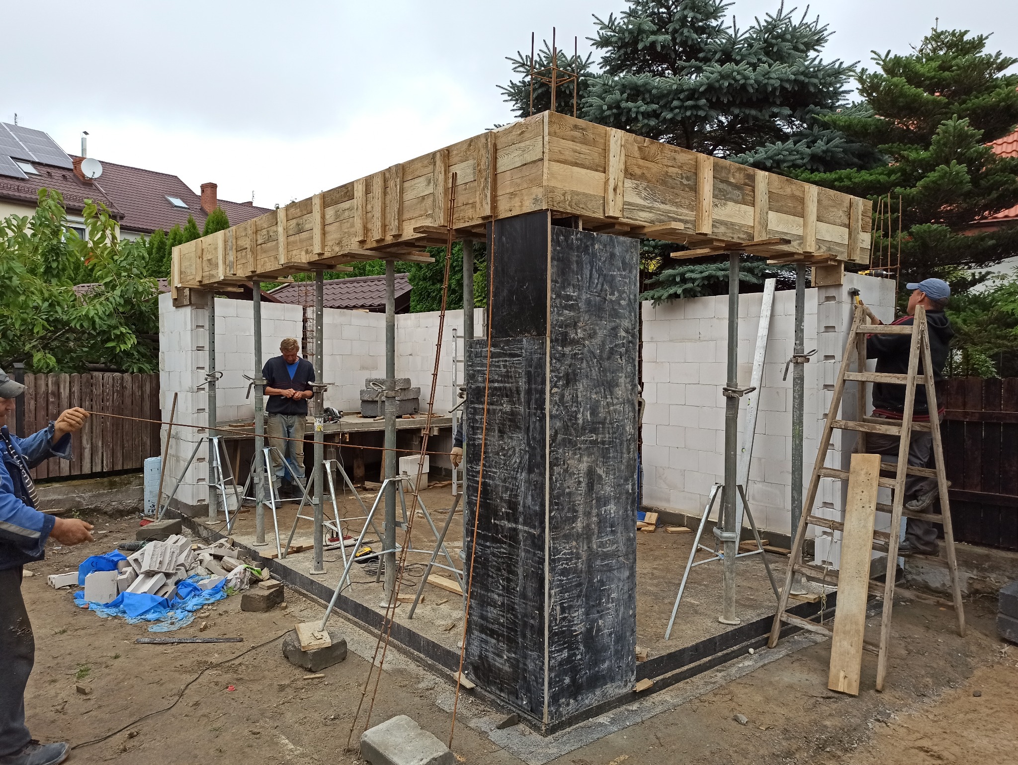 Wznoszenie konstrukcji żelbetowej: szalunek z desek podtrzymywany przez stemple, widoczni pracownicy budowlani przy pracy, bloczki betonowe i narzędzia budowlane na placu budowy.