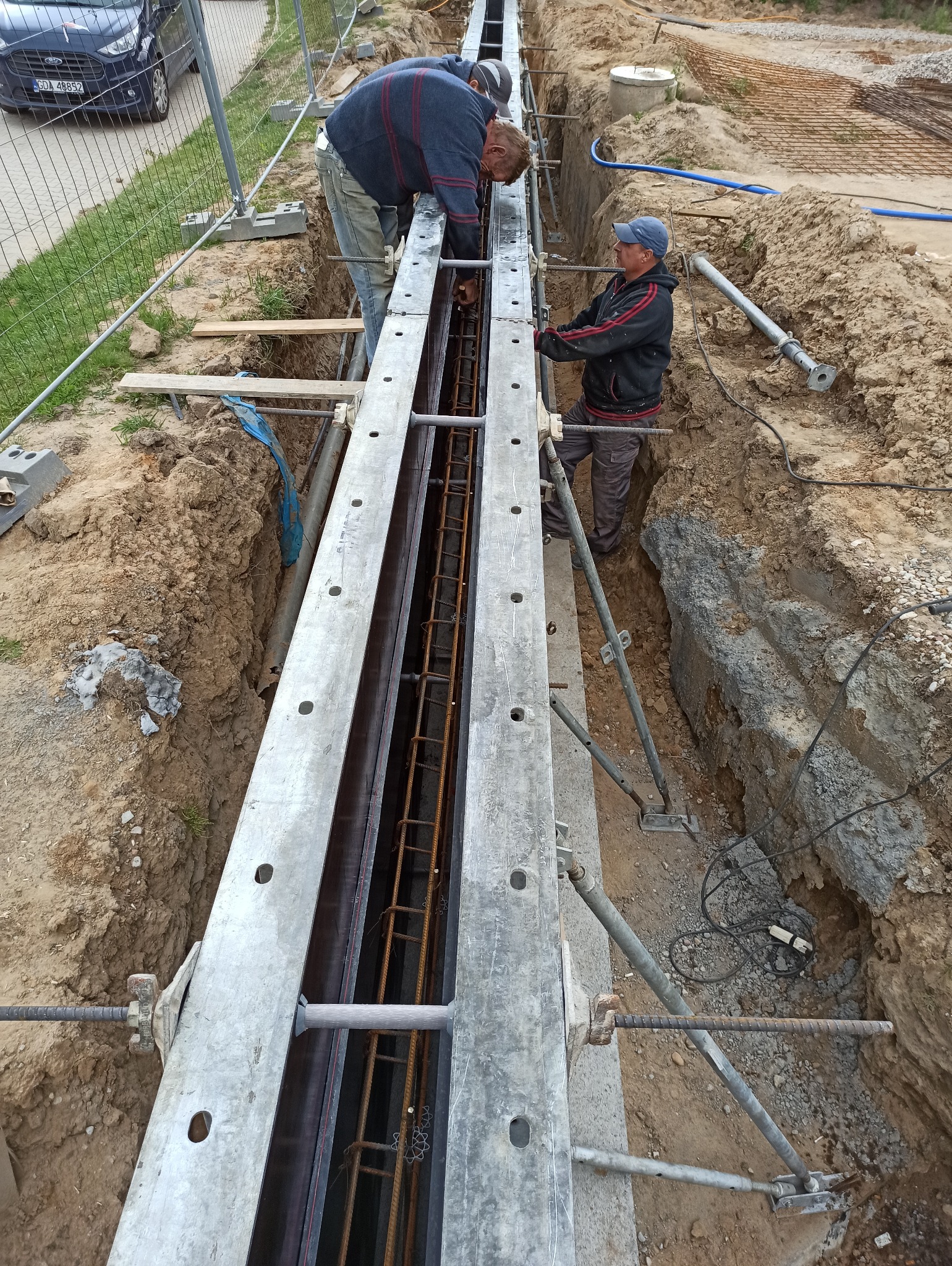 Dwóch pracowników budowlanych montuje zbrojenie w szalunku wykopu pod fundament. Widoczne metalowe elementy szalunku, stalowe pręty zbrojeniowe, ziemny wykop i elementy zabezpieczenia wykopu.