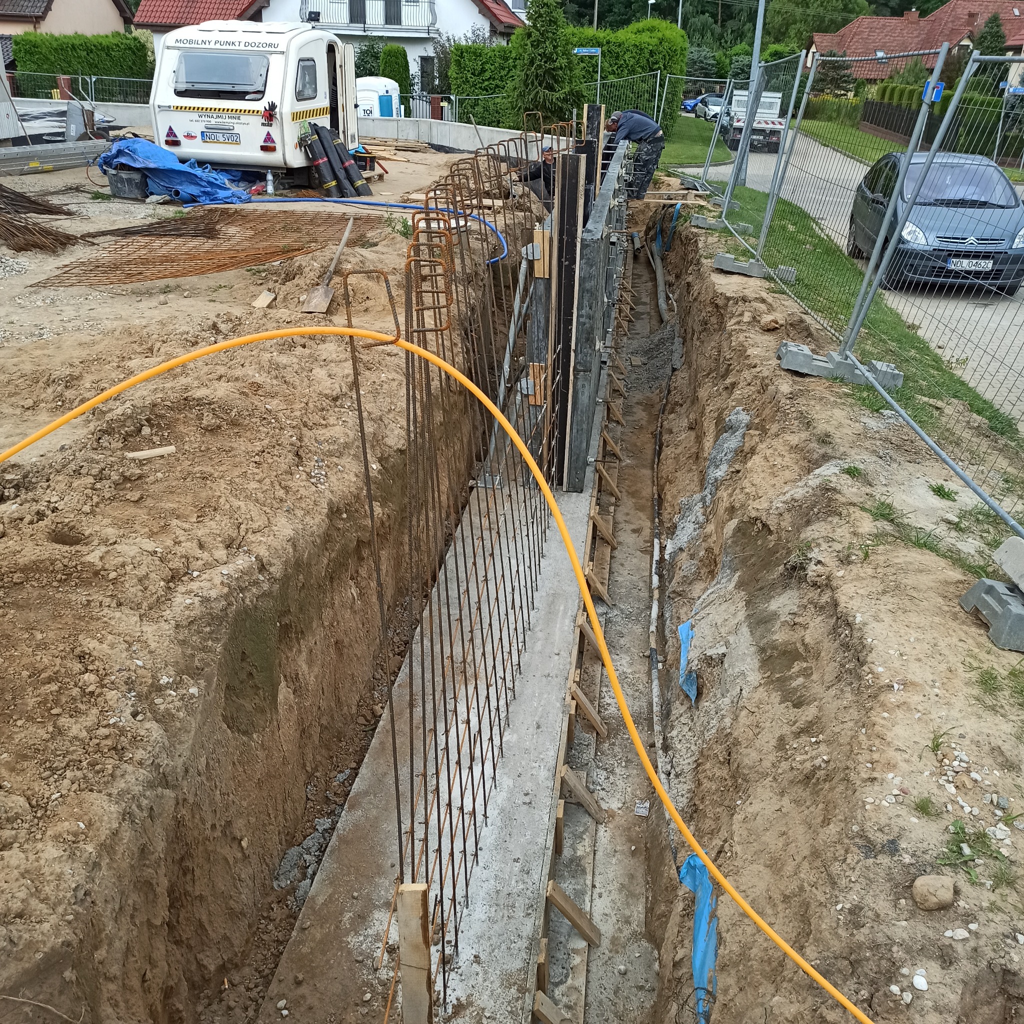 Wykop z betonowym fundamentem z wystającymi prętami zbrojeniowymi, zabezpieczony deskowaniem i żółtym kablem, widoczni pracownicy na budowie.