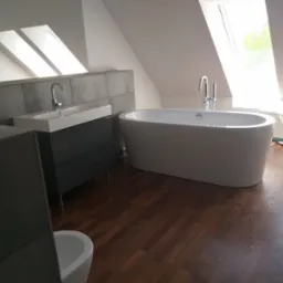 Nowoczesna łazienka na poddaszu z drewnianą podłogą, wolnostojącą wanną i umywalką nablatową na szafce w odcieniach szarości, oświetlona przez okna dachowe.