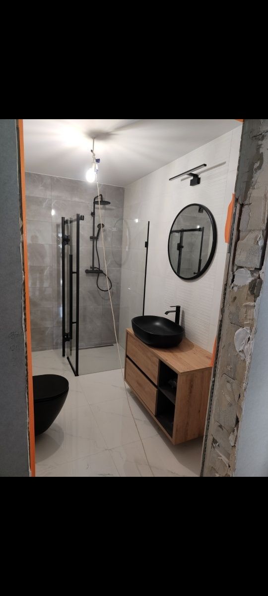 Remont łazienki w trakcie: widoczna czarna armatura, kabina prysznicowa typu walk-in, owalna umywalka na drewnianej szafce oraz niedokończone ściany.