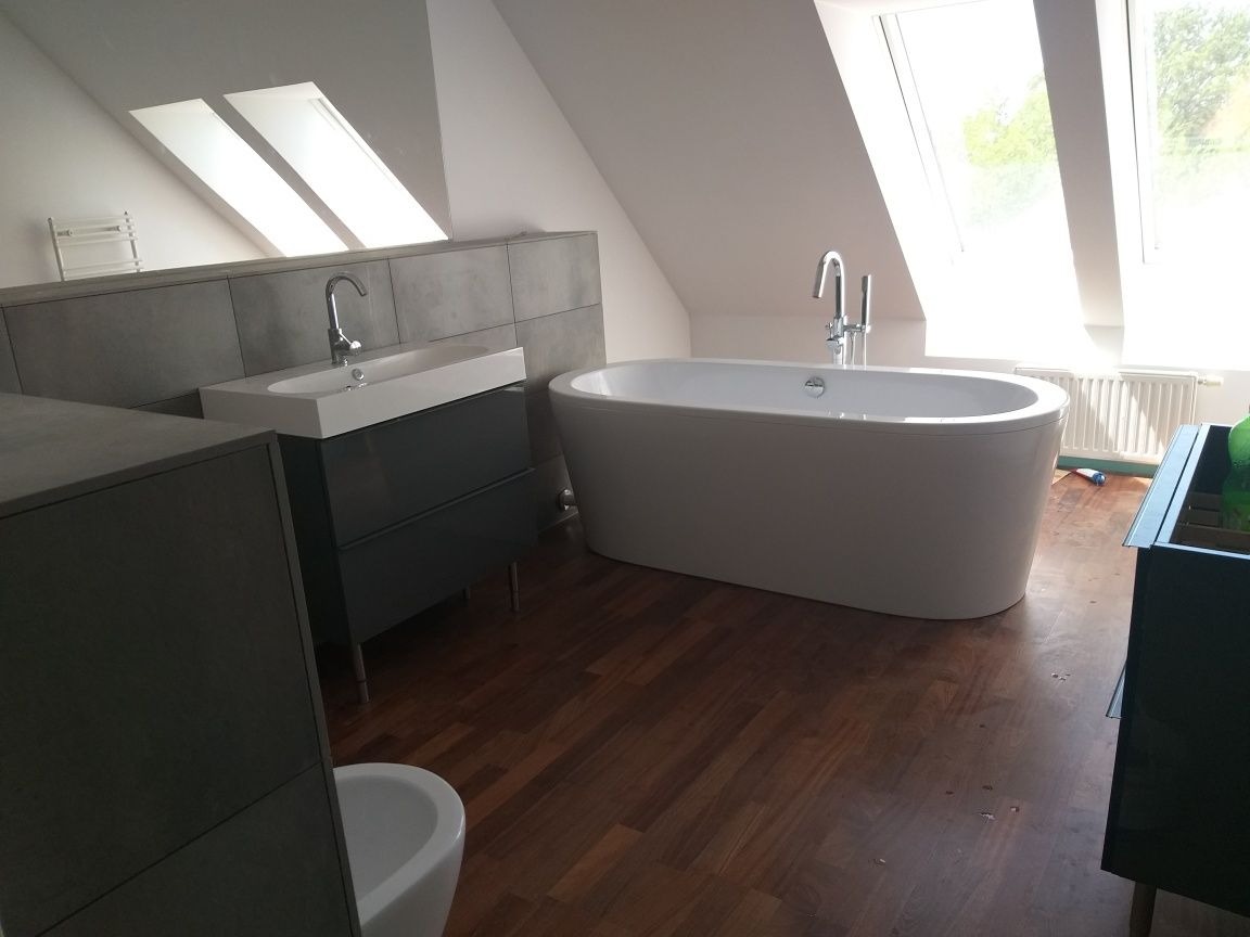 Nowoczesna łazienka na poddaszu z drewnianą podłogą, wolnostojącą wanną i umywalką nablatową na szafce w odcieniach szarości, oświetlona przez okna dachowe.