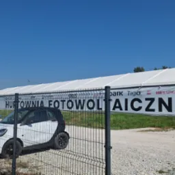 Hala magazynowa z banerem reklamującym hurtownię fotowoltaiczną, widoczna przez metalowe ogrodzenie, z zaparkowanymi samochodami.