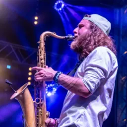 Saksofonista z długimi włosami i czapką bejsbolową gra na saksofonie na scenie oświetlonej niebieskimi światłami.