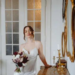 Kobieta w satynowej sukience trzyma bukiet kwiatów w dłoni, opierając się o marmurową komodę z świecznikami i stosem książek, na tle białych drzwi i luster w złoconych ramach.