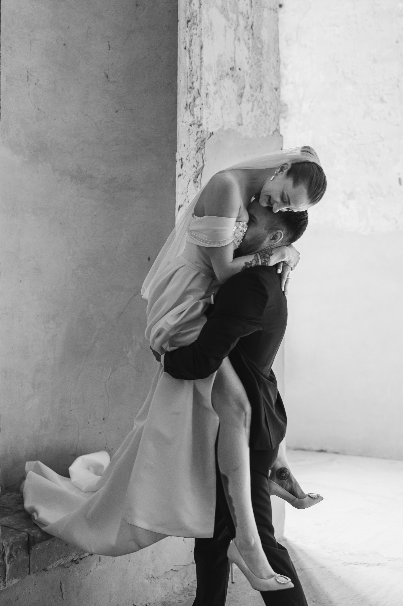 Czarno-białe zdjęcie: Pan młody niesie pannę młodą na rękach przy surowej ścianie, panna młoda w sukni z trenem i welonem, widoczny tatuaż na stopie panny młodej.