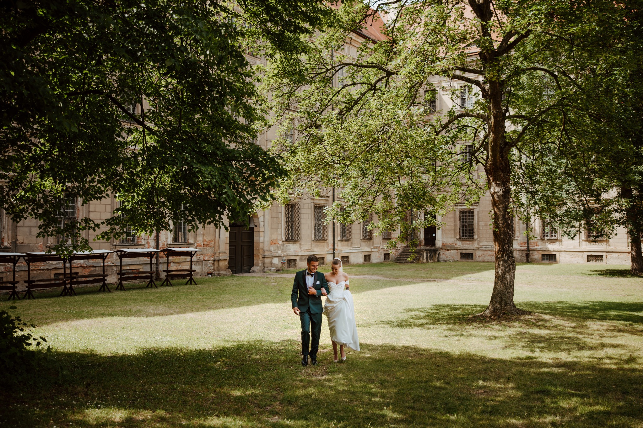Para młoda, on w butelkowej zieleni garniturze, ona w białej sukni z odsłoniętymi ramionami, spacerują po trawniku przed zabytkowym budynkiem w cieniu drzew.
