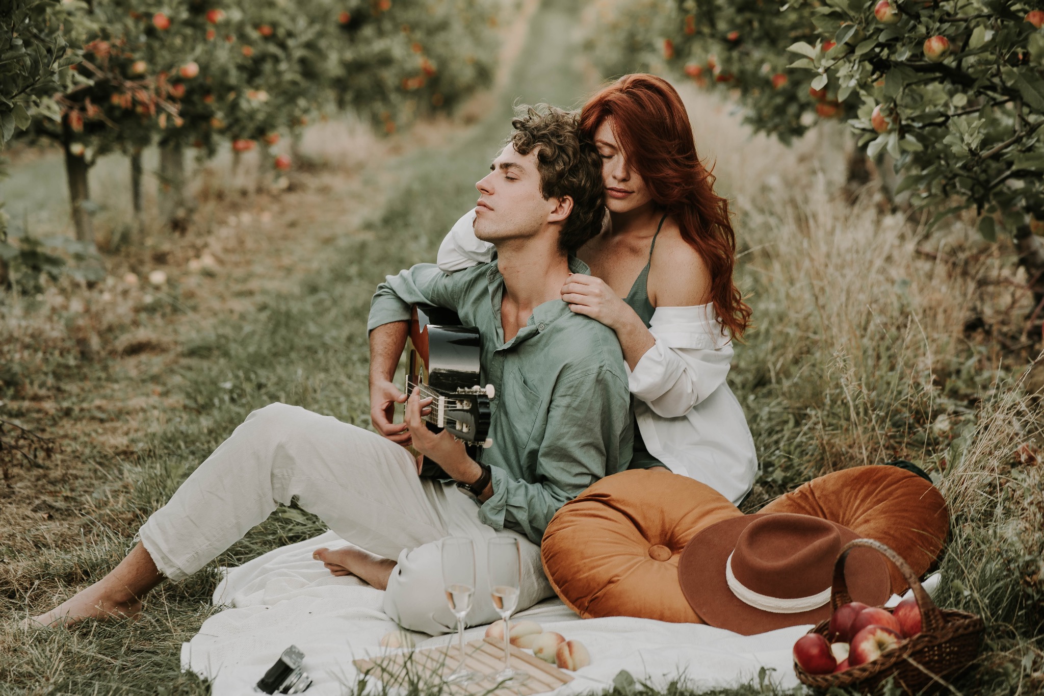 Sesja narzeczeńska we Wrocławiu: Mężczyzna gra na gitarze, obejmowany przez rudowłosą kobietę w sadzie jabłoniowym, piknik z jabłkami i szampanem.