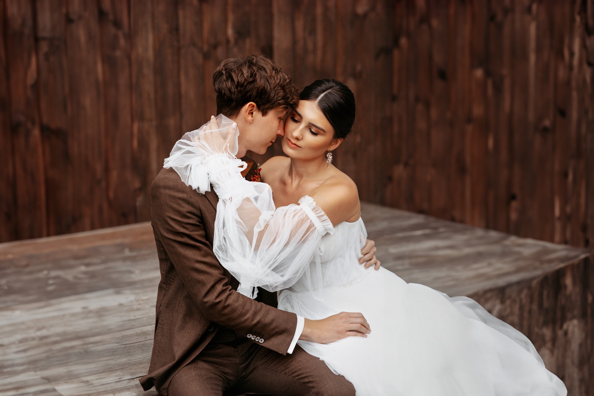 Para młoda w objęciach na drewnianym tle: panna młoda w białej sukni z bufiastymi rękawami, pan młody w brązowym garniturze.