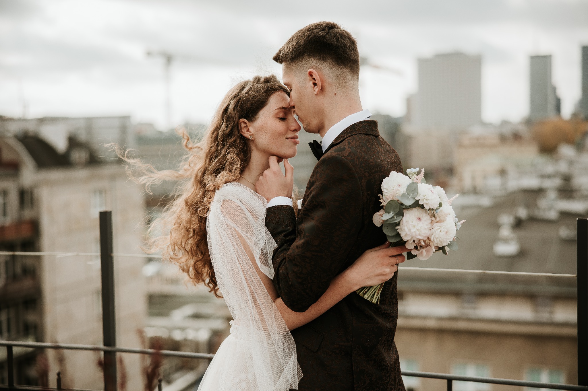 Para młoda w objęciach na tle miejskiego krajobrazu; panna młoda w zwiewnej sukience, pan młody w bordowym garniturze z bukietem w ręku.