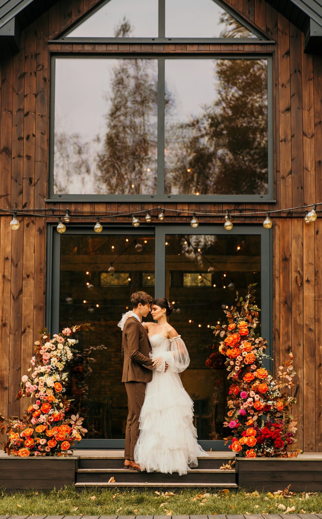 Para młoda przytulona przed drewnianym budynkiem z dużym oknem, udekorowanym girlandami kwiatowymi i żarówkami