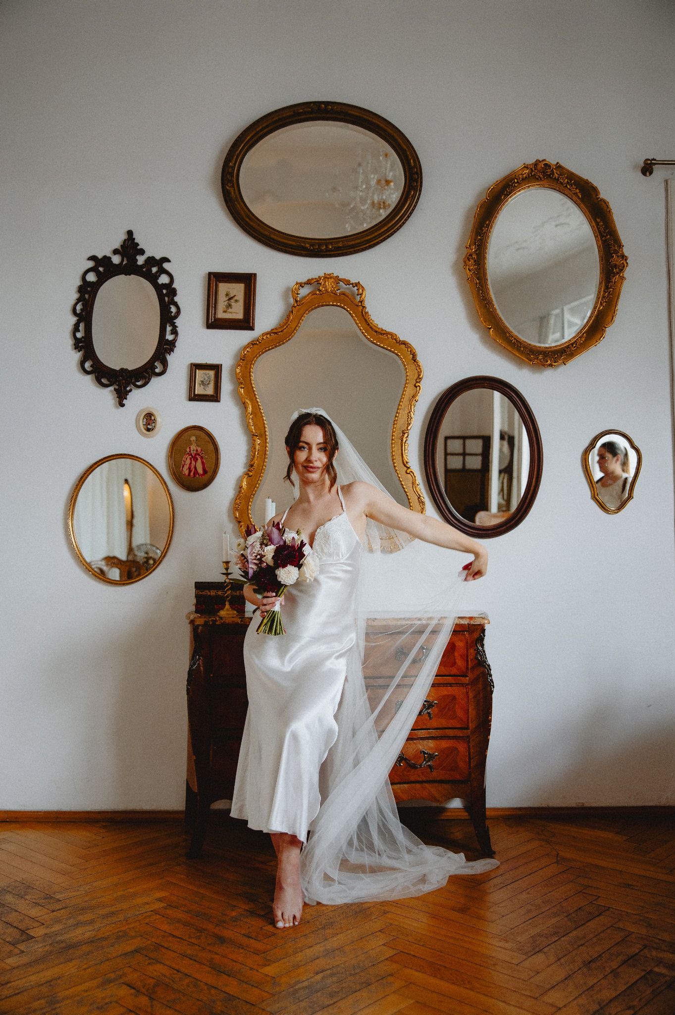 Panna młoda w satynowej sukience i welonie, trzymająca bukiet kwiatów przed komodą z lustrami w ozdobnych ramach