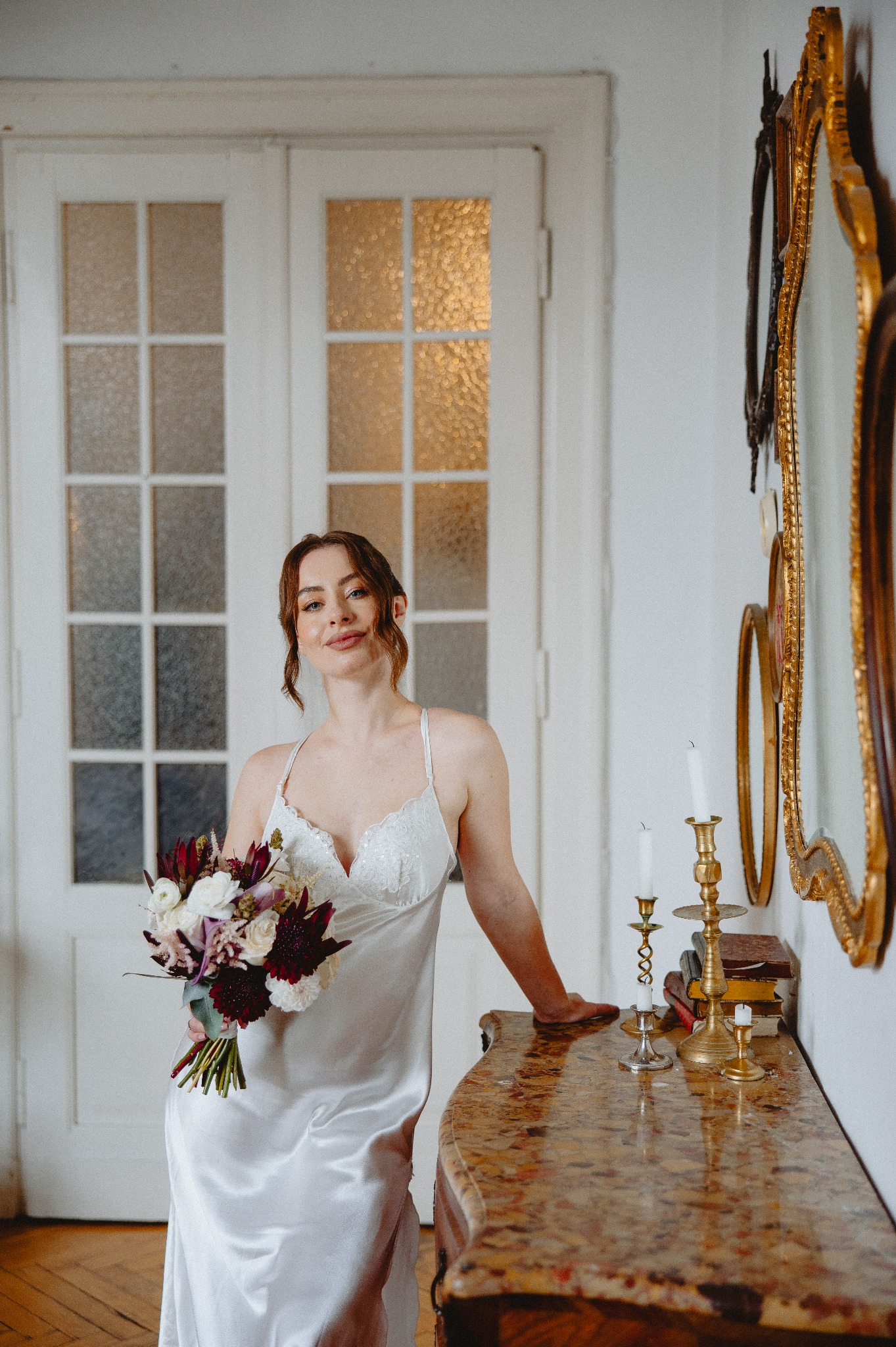 Kobieta w satynowej sukience trzyma bukiet kwiatów w dłoni, opierając się o marmurową komodę z świecznikami i stosem książek, na tle białych drzwi i luster w złoconych ramach.