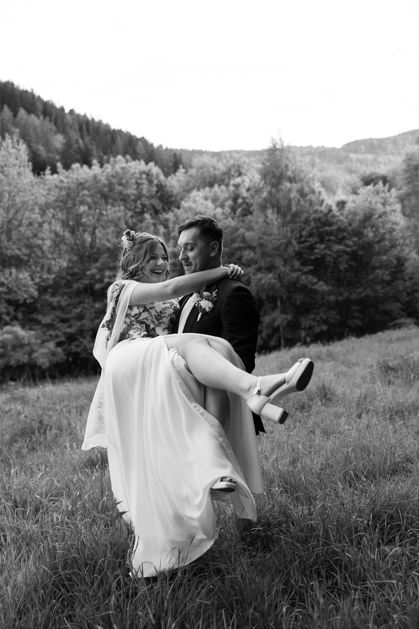 Czarno-białe zdjęcie: Uśmiechnięty pan młody trzyma pannę młodą na rękach na tle łąki i drzew. Panna młoda w sukni z haftem i kwiatami we włosach.