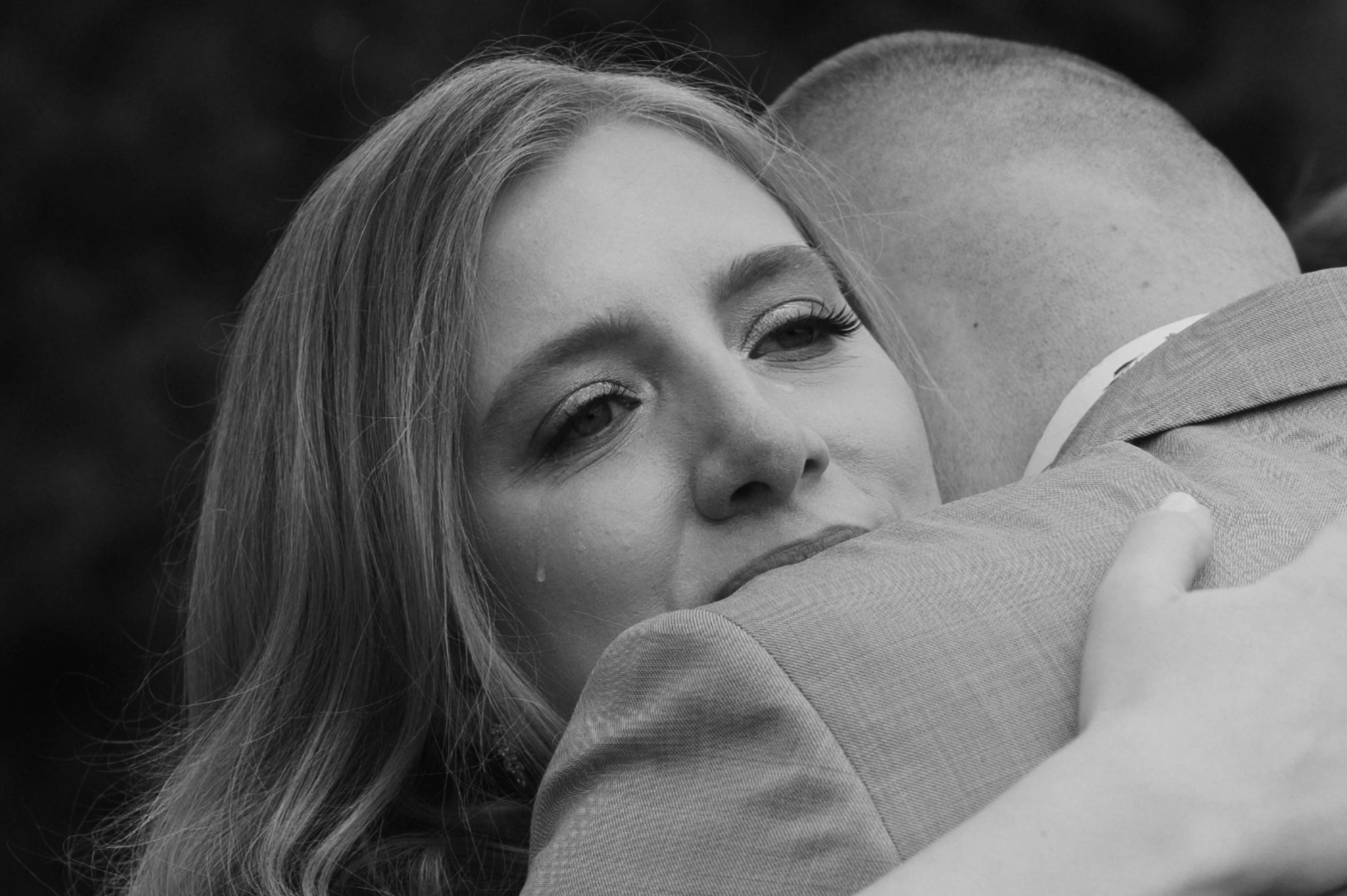 Czarno-białe zbliżenie kobiety z łzą w oku, obejmującej mężczyznę w garniturze. Ujęcie z uroczystości, skupione na emocjach i detalu twarzy.