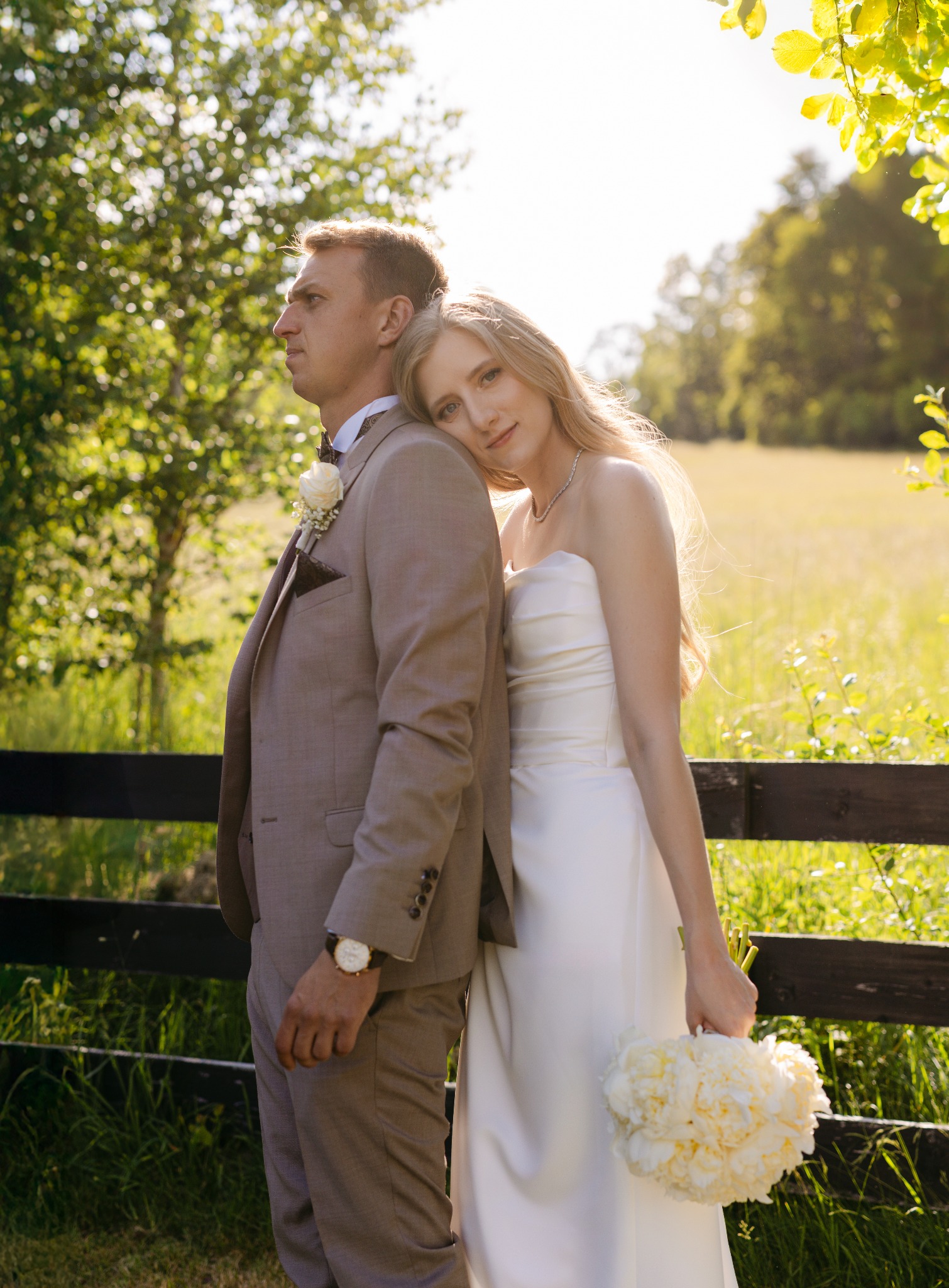 Para młoda w plenerze: panna młoda opiera głowę na ramieniu pana młodego, trzyma bukiet piwonii. W tle zielone drzewa i drewniany płot.