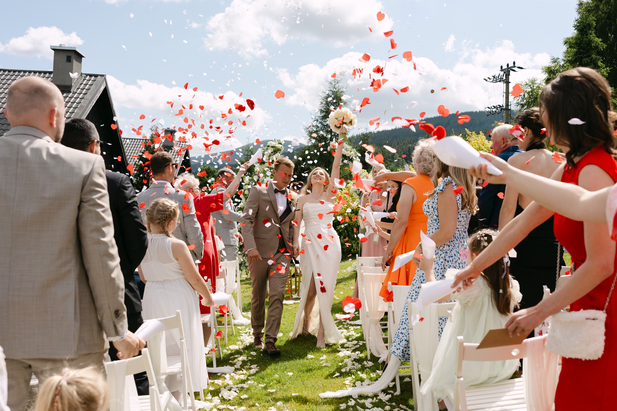 Para młoda w otoczeniu gości weselnych, obsypywana konfetti w kształcie serc na tle zieleni i gór. Panna młoda trzyma bukiet.