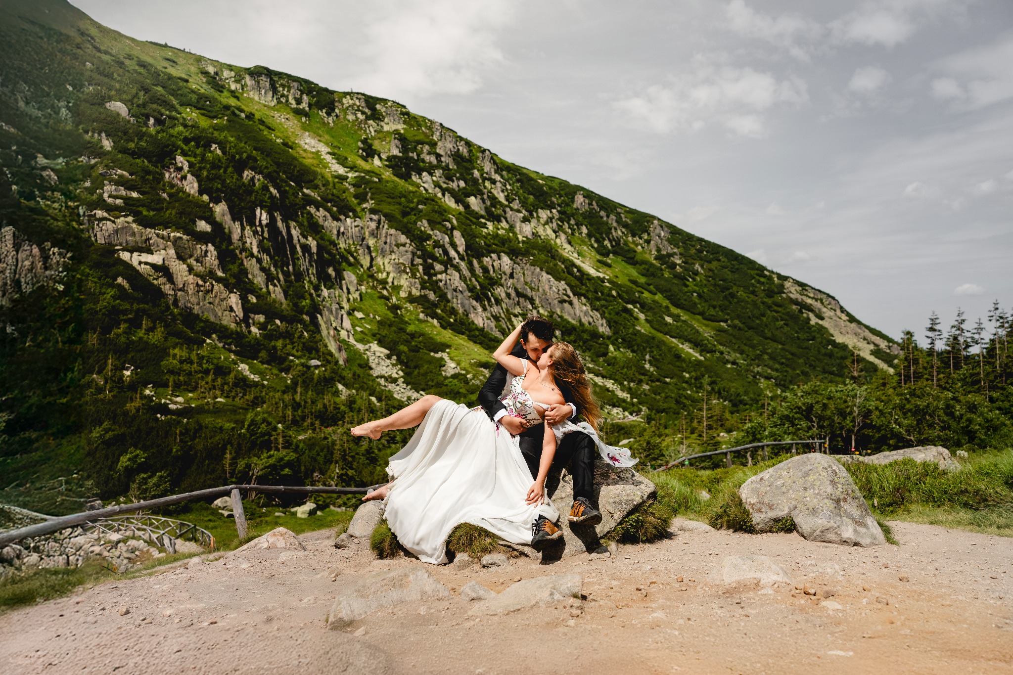 Para młoda w górach: Panna Młoda w białej sukni i kwiatowym gorsecie, Pan Młody w czarnym garniturze, siedzą na kamieniu na tle zielonego zbocza.