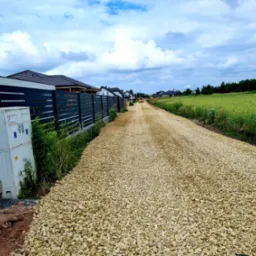 Droga wysypana jasnym kruszywem, obok nowoczesne ogrodzenie z elementami betonu i metalu, w tle zabudowa jednorodzinna i pole uprawne pod pochmurnym niebem.
