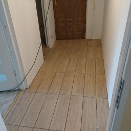 Zient-bud - Ułożone płytki podłogowe imitujące drewno w przedpokoju, widoczne niedokończone prace elektryczne i surowe wykończenie ścian.