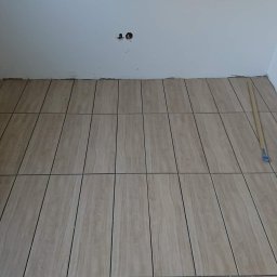 Zient-bud - Ułożone podłużne płytki podłogowe imitujące drewno, z widoczną fugą, na ścianie płytki z motywem kwiatowym i niedokończone gniazdko elektryczne, widoczne narzędzie do rozprowadzania kleju.