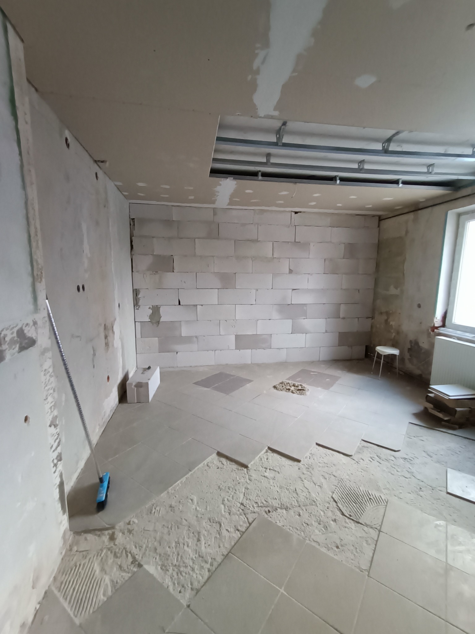 Wnętrze w trakcie prac: częściowo ułożone płytki podłogowe, ściana z betonu komórkowego, surowy sufit z widoczną konstrukcją metalową i fragmentami płyty karton-gips, oraz listwa przypodłogowa przy...