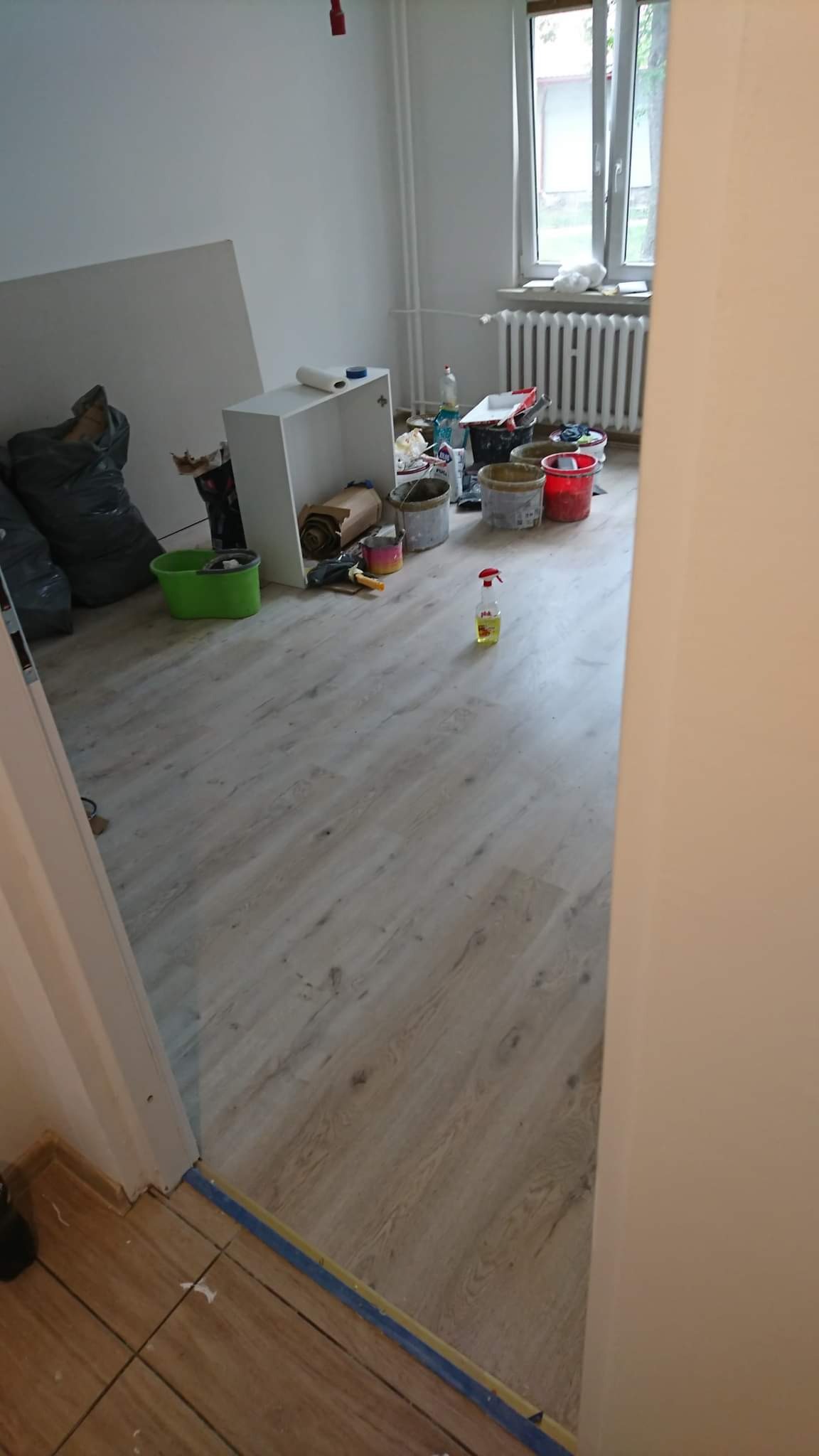 Pokój w trakcie prac, widoczne wiadra z farbą, pędzle i akcesoria malarskie, zabezpieczona taśmą podłoga, ślady po pracach na podłodze.