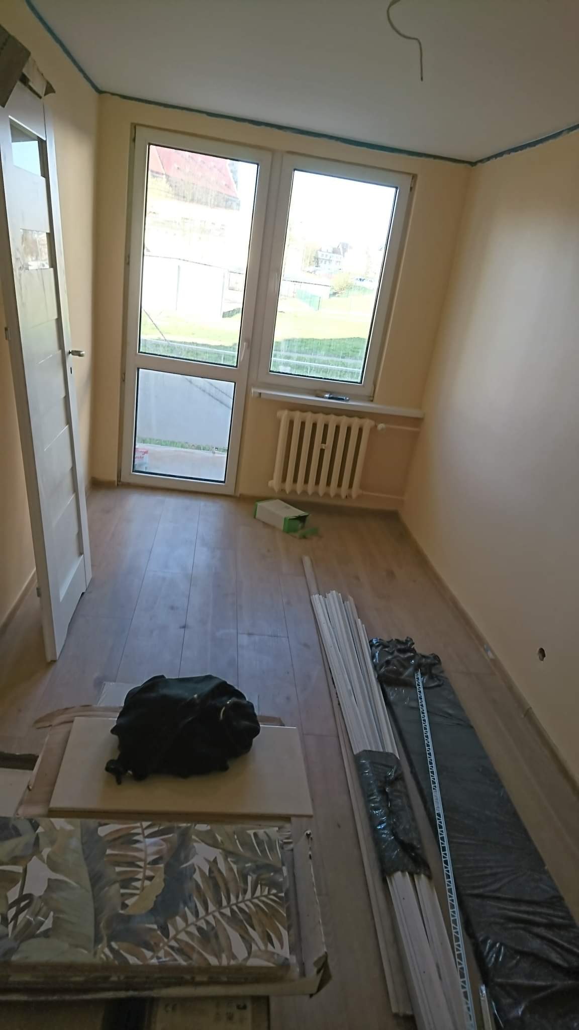 Pokój w trakcie prac, widoczne materiały budowlane, listwy przypodłogowe, płytki z motywem roślinnym, oklejone taśmą malarską ściany i sufit, oraz grzejnik pod oknem.