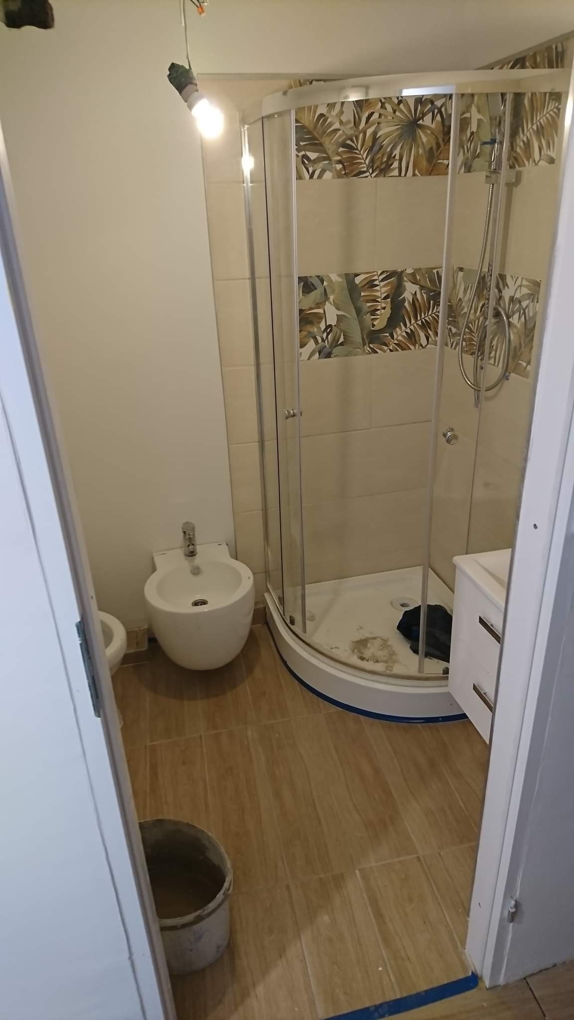 Nowoczesna łazienka z kabiną prysznicową z motywem liści na płytkach, bidetem i wiadrem z zaprawą, oświetlona żarówką na kablu.