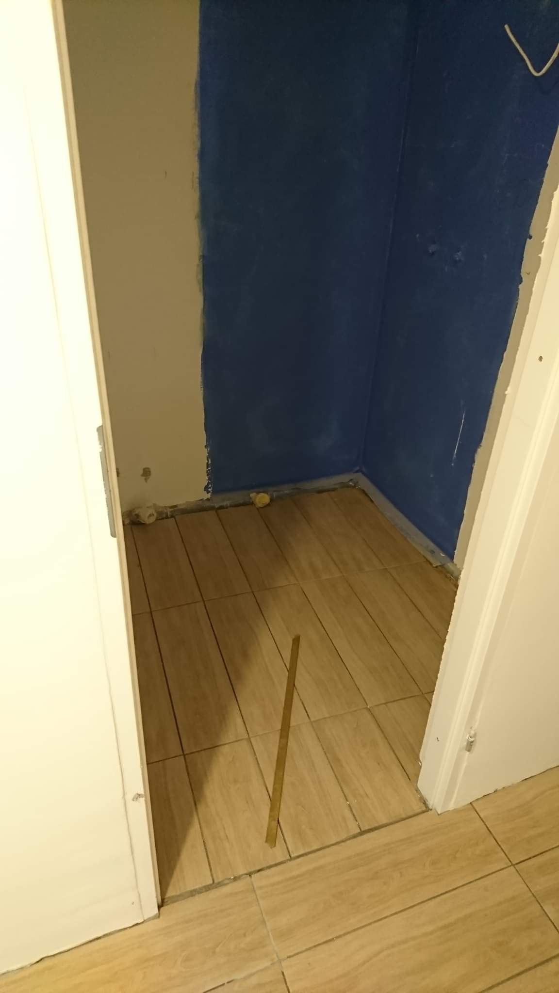 Wnęka w trakcie prac: częściowo pomalowana na niebiesko ściana, fragment ściany w kolorze kremowym, ułożona podłoga z płytek imitujących drewno, widoczna listwa przypodłogowa i rurki przy podłodze.