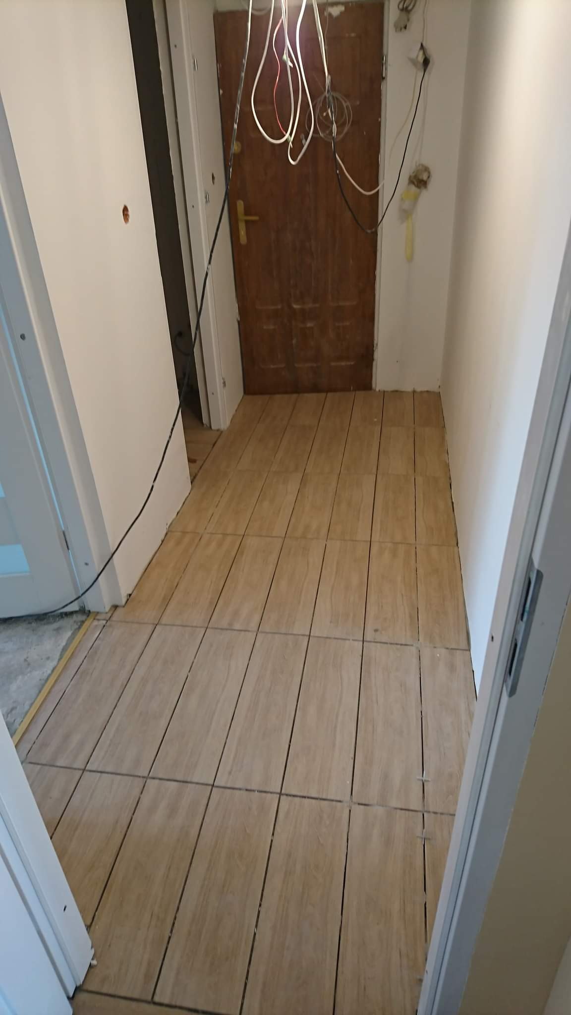 Ułożone płytki podłogowe imitujące drewno w przedpokoju, widoczne niedokończone prace elektryczne i surowe wykończenie ścian.
