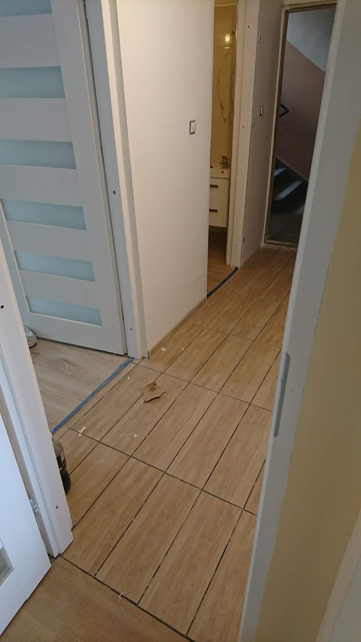 Korytarz z ułożonymi płytkami imitującymi drewno, zabezpieczony niebieską taśmą malarską przy ścianach, widoczne drzwi i fragment łazienki w tle.