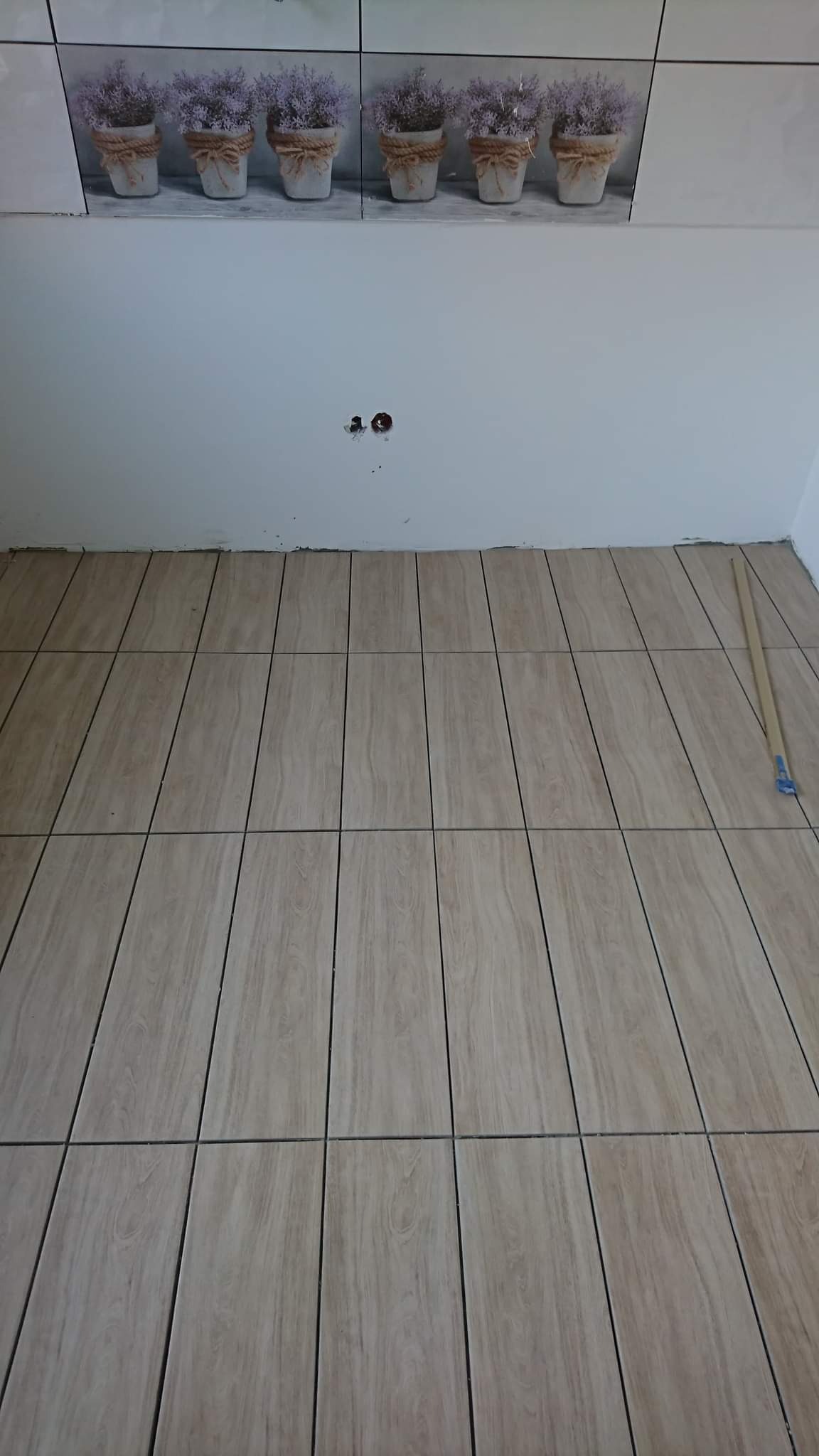 Ułożone podłużne płytki podłogowe imitujące drewno, z widoczną fugą, na ścianie płytki z motywem kwiatowym i niedokończone gniazdko elektryczne, widoczne narzędzie do rozprowadzania kleju.