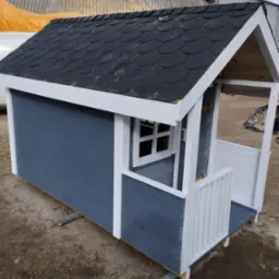 Niebiesko-biały domek dla dzieci z czarnym dachem z gontu bitumicznego, ustawiony na rurach na ziemi.