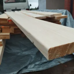 Stos jasnych, świeżo przetartych desek ułożonych na folii ochronnej, widoczne ślady po cięciu i naturalny rysunek drewna, w tle samochód dostawczy i fragment budynku.