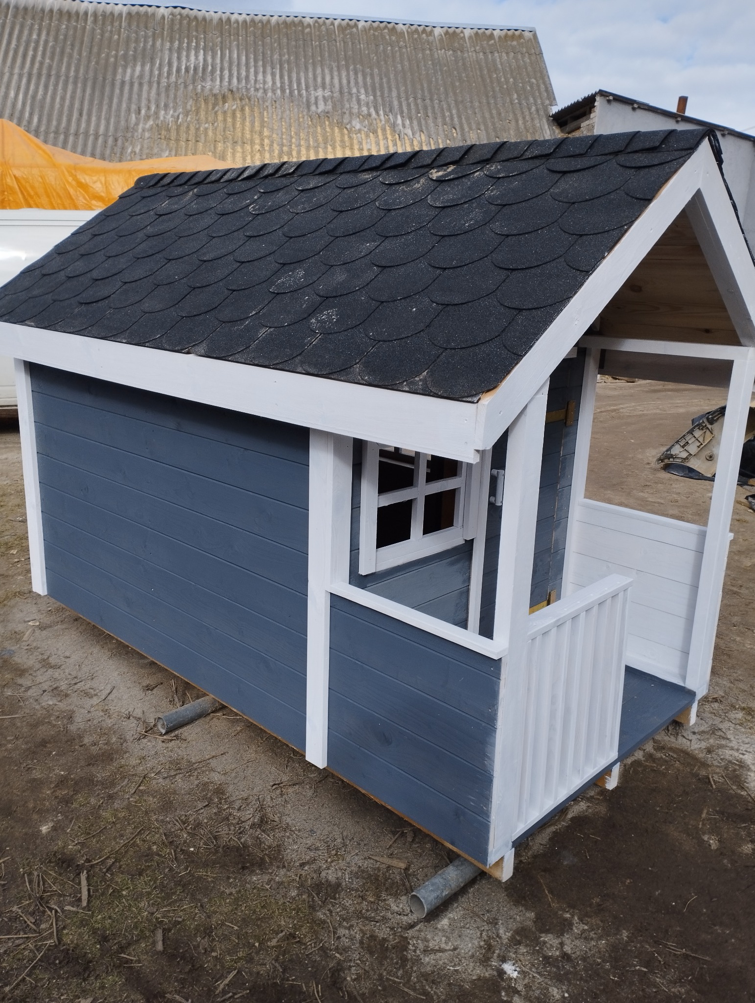 Niebiesko-biały domek dla dzieci z czarnym dachem z gontu bitumicznego, ustawiony na rurach na ziemi.