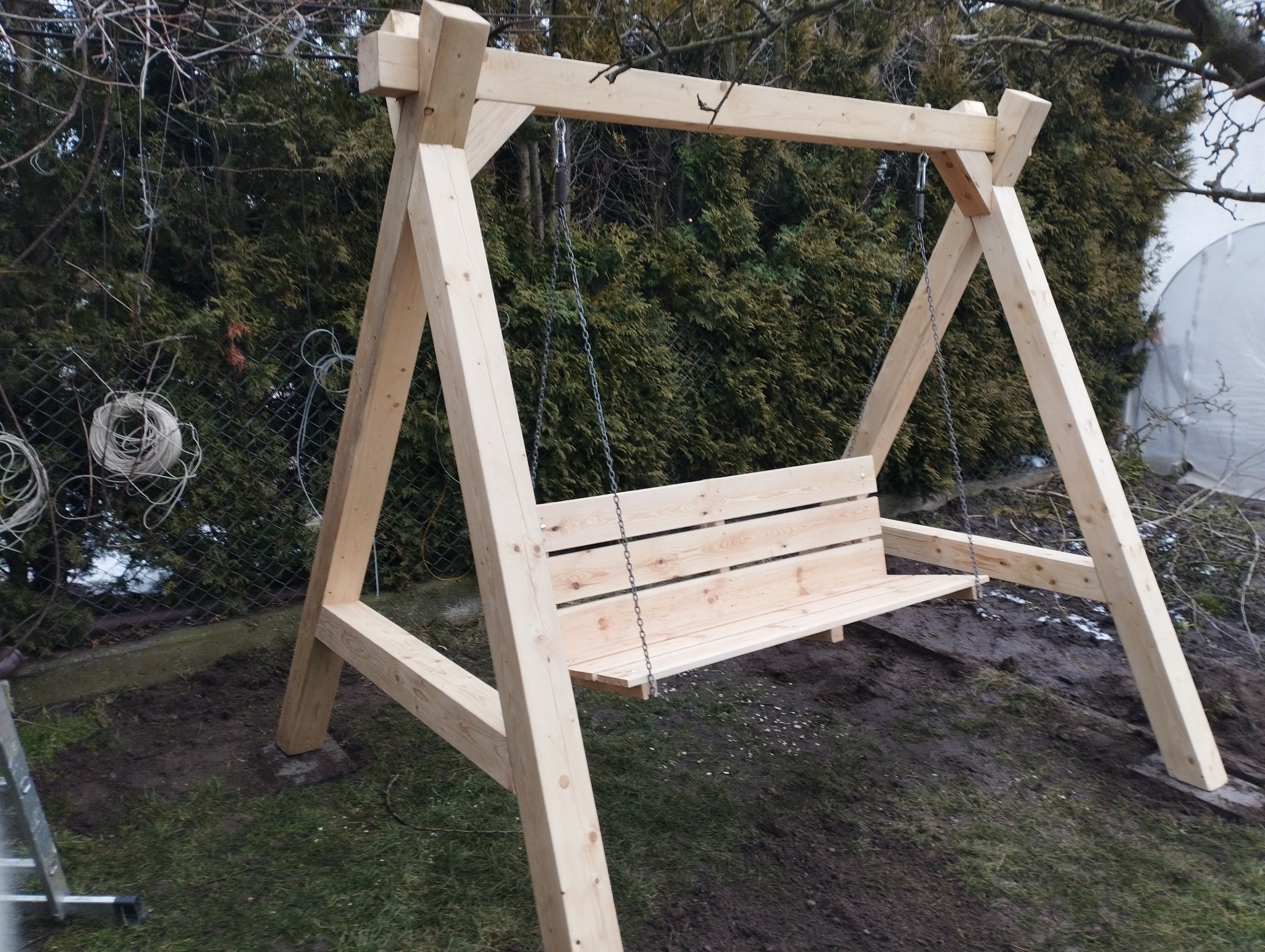 Nowa, drewniana huśtawka ogrodowa na metalowych łańcuchach, z surowego jasnego drewna, ustawiona w ogrodzie na trawie, z widocznymi krzewami w tle.
