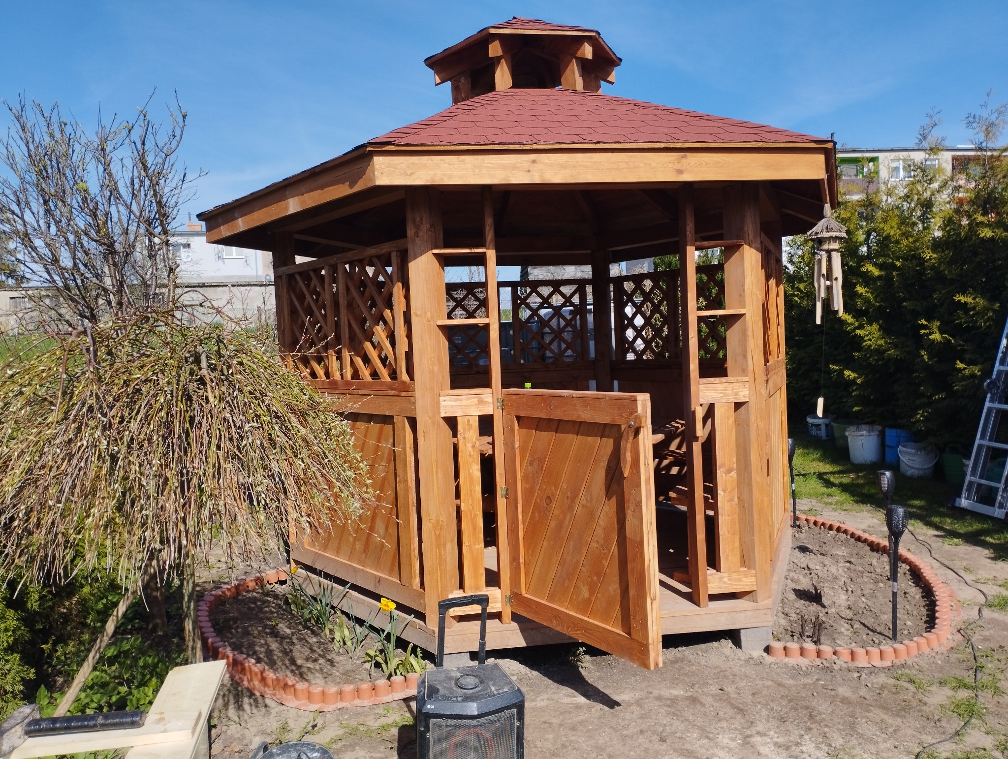 Drewniana altana z czerwoną dachówką, otwartymi drzwiami i kratownicowymi ścianami, otoczona rabatą z terakoty i młodymi roślinami, z głośnikiem bluetooth na pierwszym planie.