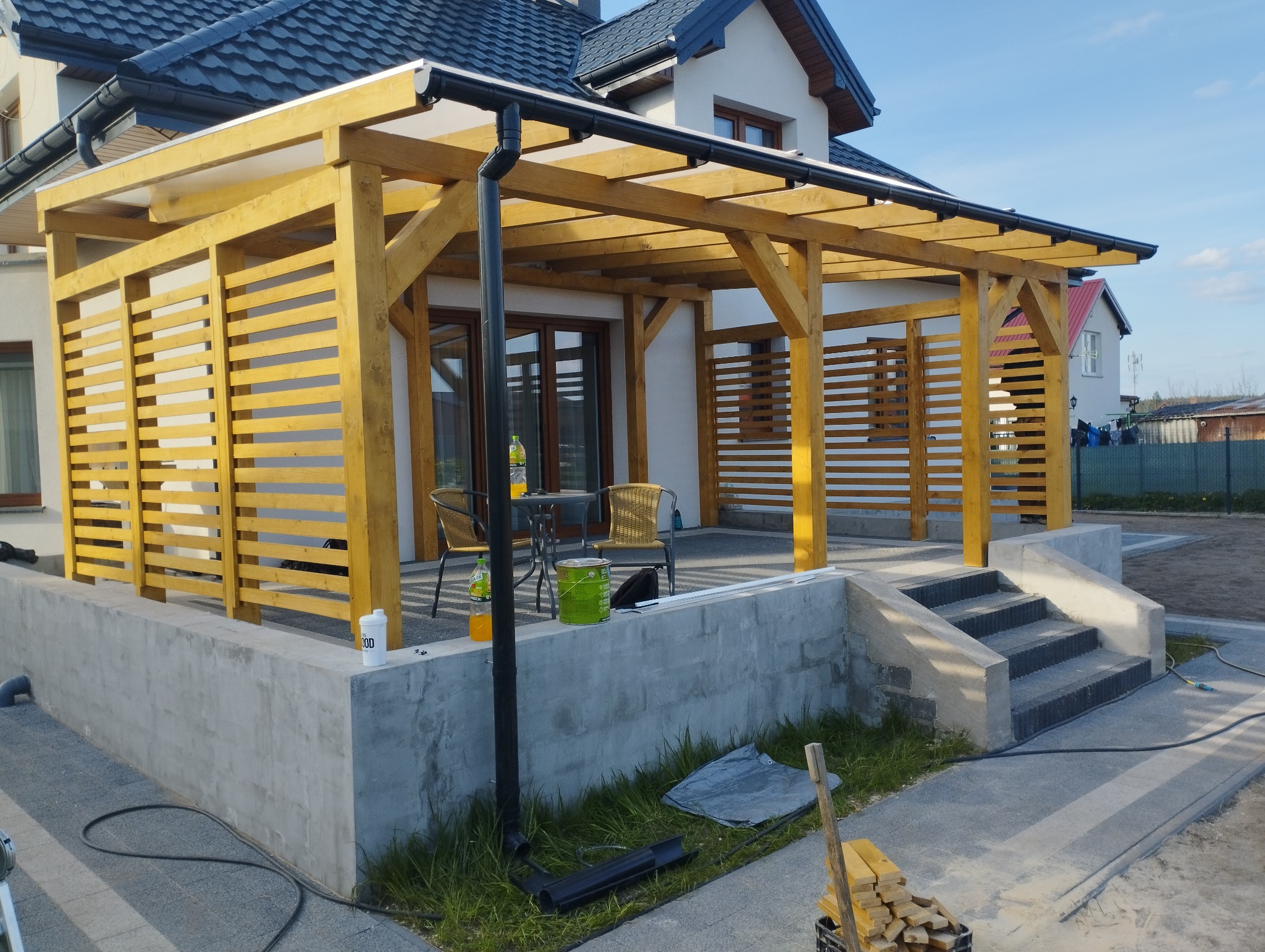 Nowoczesna altana z drewna o jasnym odcieniu, z ażurowymi ścianami i przezroczystym zadaszeniem, przylegająca do domu z widocznymi schodami wejściowymi i elementami wykończeniowymi w trakcie prac.