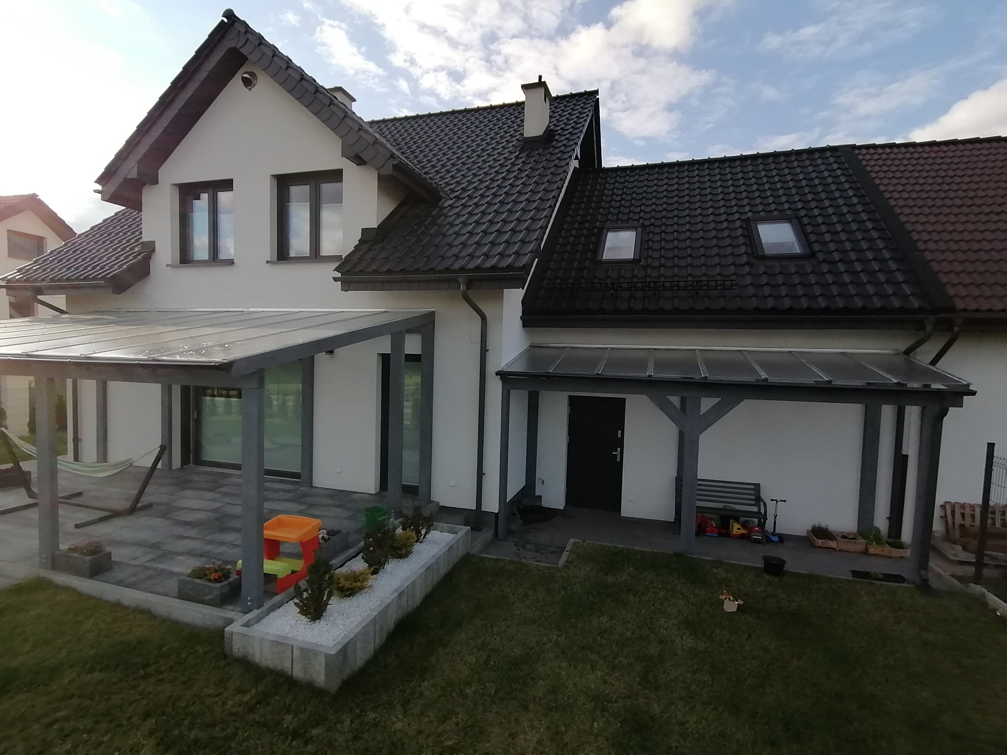 Nowoczesny dom z białymi ścianami i ciemną dachówką, zadaszony taras z szarą kostką brukową, widoczne elementy zagospodarowania ogrodu: rabata z białym grysem, trawnik, hamak i zabawki dziecięce.