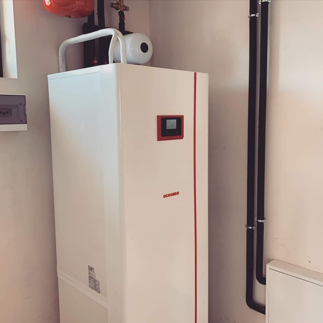 Biała jednostka grzewcza Ochsner z czerwonym akcentem i wyświetlaczem, zainstalowana w pomieszczeniu z rurami i skrzynką elektryczną.