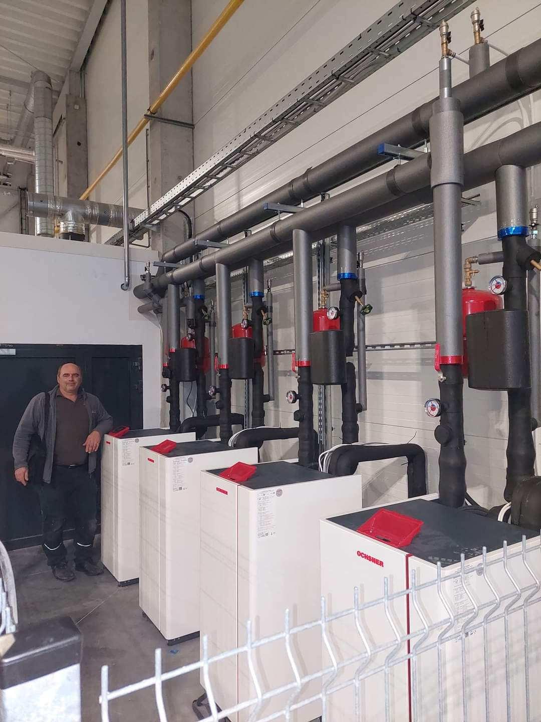 Instalacja czterech białych jednostek grzewczych Ochsner z czerwonymi elementami, widoczne rury w izolacji, pracownik oparty o jedną z jednostek, metalowa siatka na pierwszym planie.